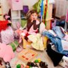 Überforderte Mädchen sitzt in chaotischem Kinderzimmer zwischen Kleidung und Spielzeug